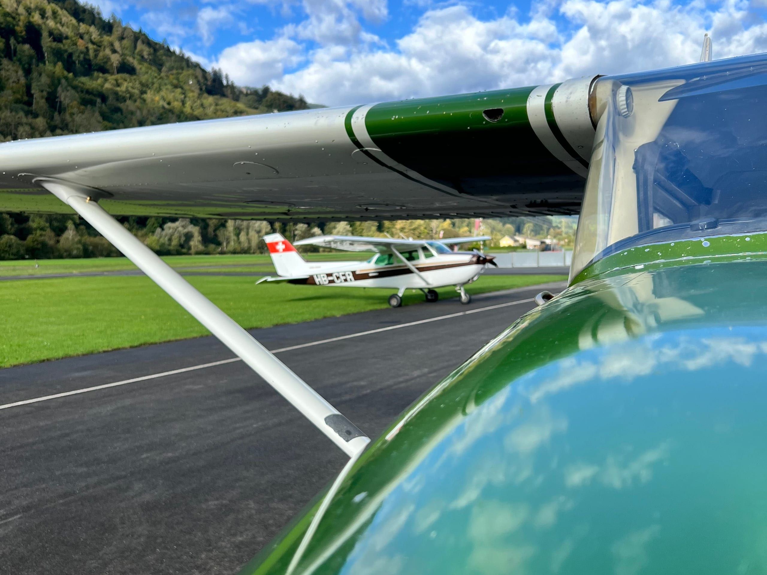 At Bad Ragaz airfield: looking under the wing of the green HB-CEU towards HB-CFR, forest and clouds behind