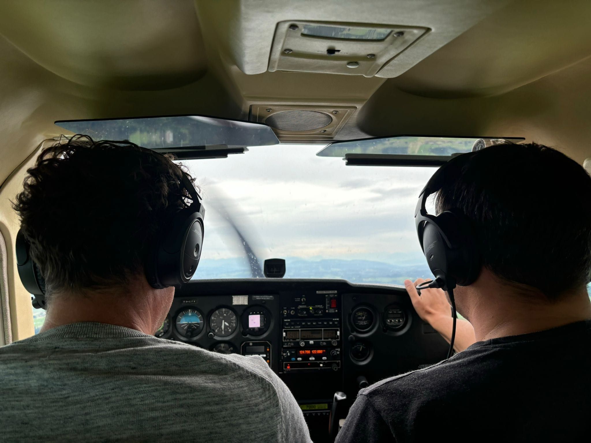 In the cockpit from behind: two people with headsets, instruments and propeller ahead, land under clouds outside